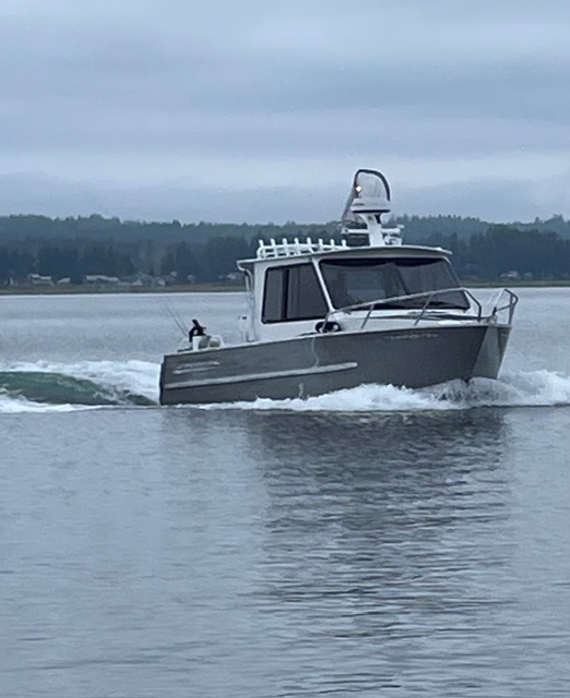 Kwigwis Adventures fishing charter boat viewed head-on from the water, displaying the boat in motion with water splashing around the hull. The boat features a white cabin with large front windows and is equipped with radar and communication equipment on top, set against a backdrop of calm water and distant forested shoreline under overcast skies.